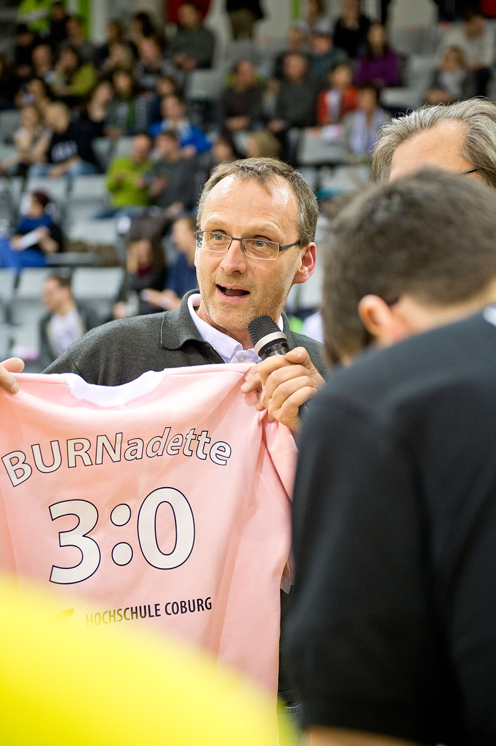 Ein Mann mit einem rosa T-Shirt mit der Aufschrift „BURNadette 3:0“ spricht in ein Mikrofon. Umgeben von einer geschäftigen Menschenmenge bei einem scheinbar sportlichen Ereignis vertritt er stolz die Hochschule Coburg in der lebhaften Atmosphäre.