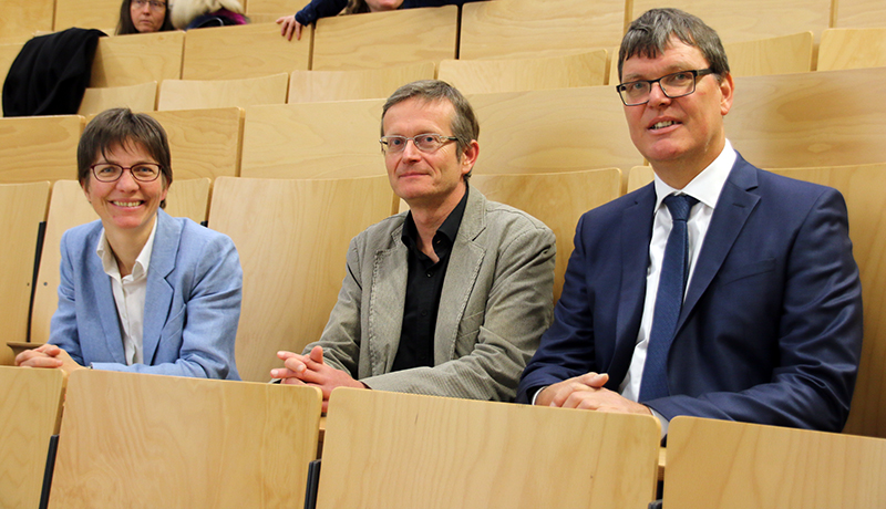 Drei Personen sitzen in einem Hörsaal der Hochschule Coburg. Die Person links trägt eine hellblaue Jacke, die mittlere eine graue und die rechte einen marineblauen Anzug. Sie blicken nach vorne und sitzen bequem auf hellen Holzstühlen.