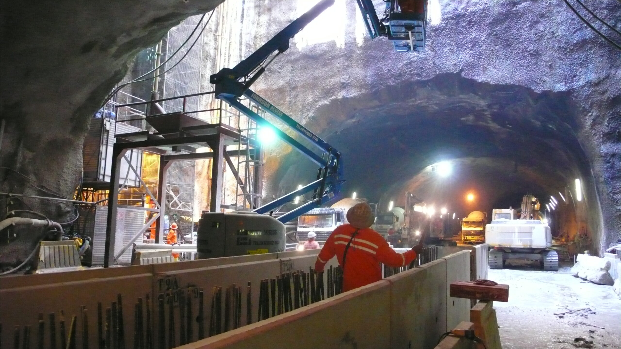 Arbeiter bauen mit Maschinen einen Tunnel, der von künstlichem Licht beleuchtet wird und an die fortgeschrittenen Ingenieurprojekte erinnert, die an der Hochschule Coburg gelehrt werden. Eine Person in orangefarbener Uniform steuert einen großen Aufzug, während in dem unterirdischen Gang Fahrzeuge und Geräte zu sehen sind.