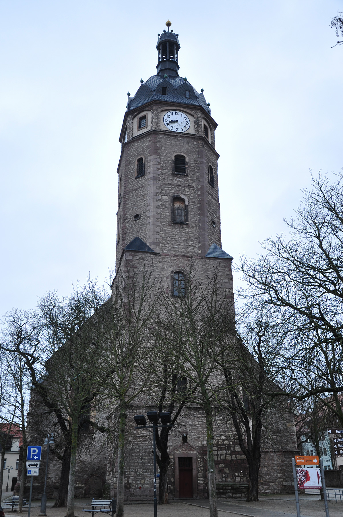 Ein hoher, historischer Uhrenturm aus Stein ragt an der Hochschule Coburg in den Himmel, umgeben von kahlen Bäumen. Der Turm verfügt über ein Spitzdach mit kleinen Dachgauben und einem auf der Vorderseite sichtbaren Zifferblatt.