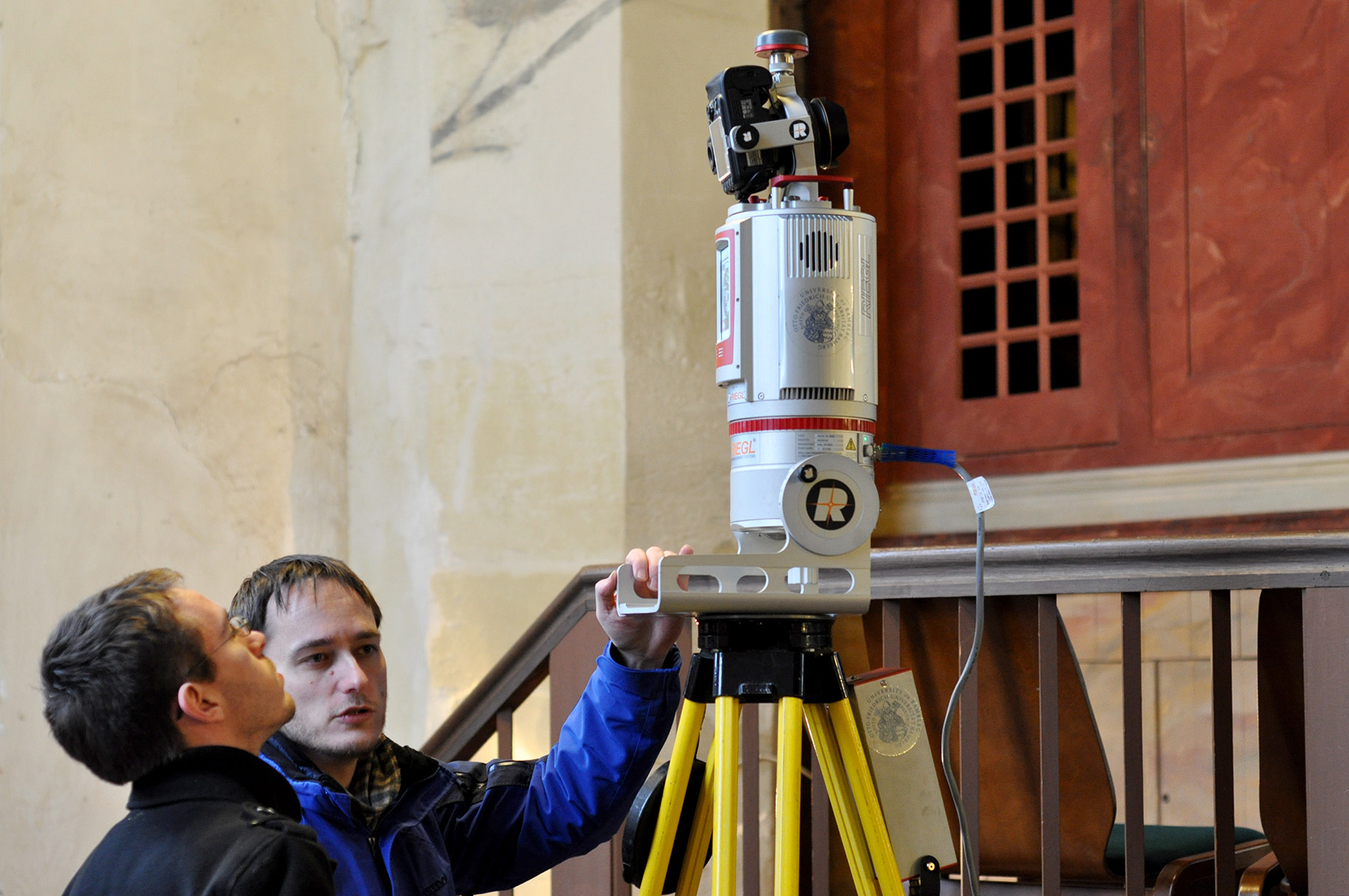 Zwei Personen der Hochschule Coburg untersuchen ein auf einem Stativ montiertes Laserscanning-Gerät. Im Innenbereich in der Nähe einer Treppe und eines Holzgeländers positioniert, justiert eine Person das Gerät, während die andere es genau beobachtet. Die Umgebung strahlt den Charme und die Eleganz eines historischen Gebäudes aus.