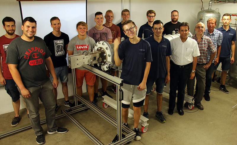 Eine Gruppe von sechzehn Männern, einige in Freizeitkleidung, steht drinnen um ein großes mechanisches Gerät auf einem Metallrahmen herum. Die Szene spielt in einer Werkstatt oder einem Labor der Hochschule Coburg.