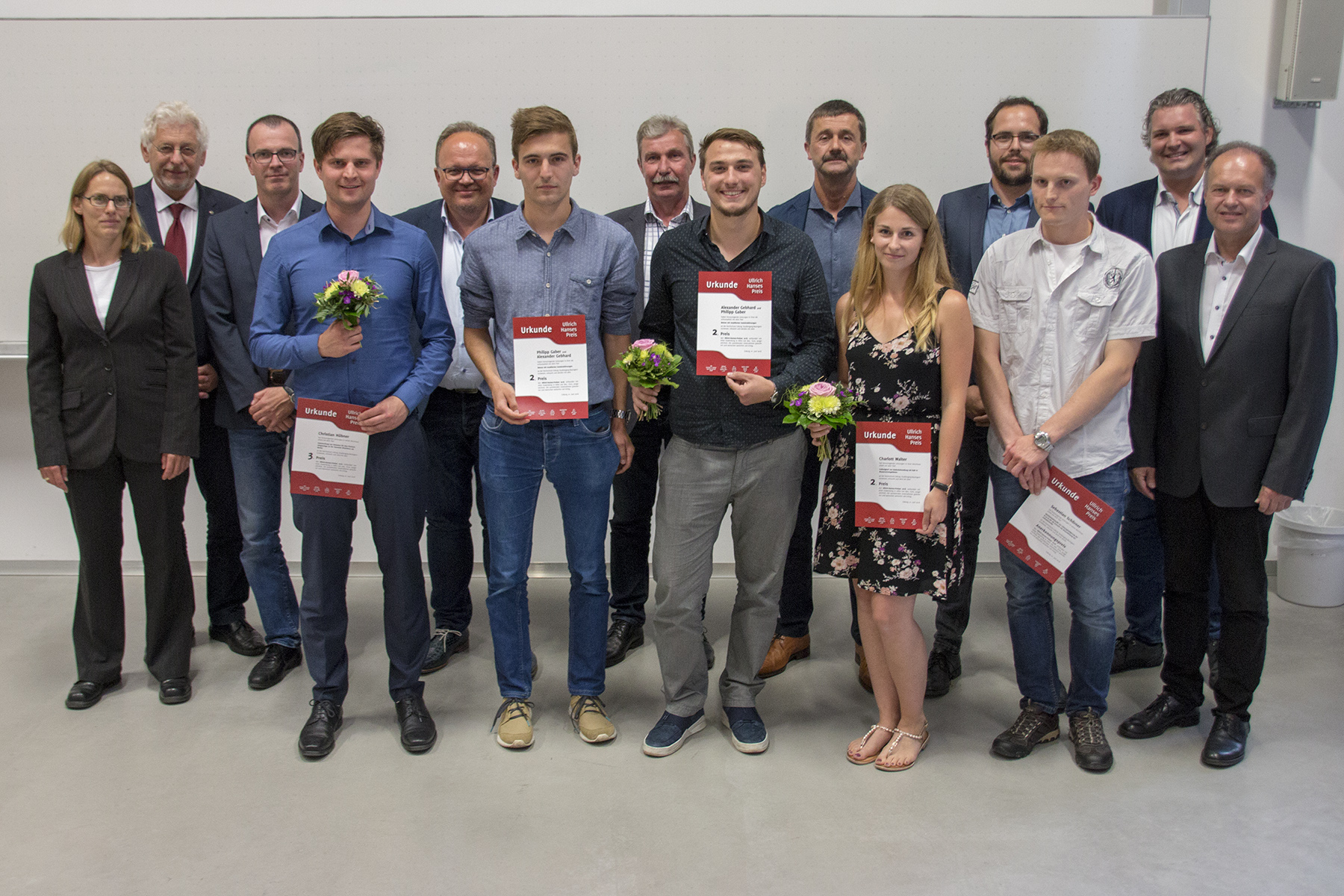 Eine Gruppe von Menschen posiert im Innenbereich der Hochschule Coburg. Vier Personen in der ersten Reihe halten Zertifikate und kleine Blumensträuße, während andere in formeller Kleidung hinter ihnen stehen. Die Szene lässt auf eine Anerkennungs- oder Preisverleihung an dieser angesehenen Institution schließen.