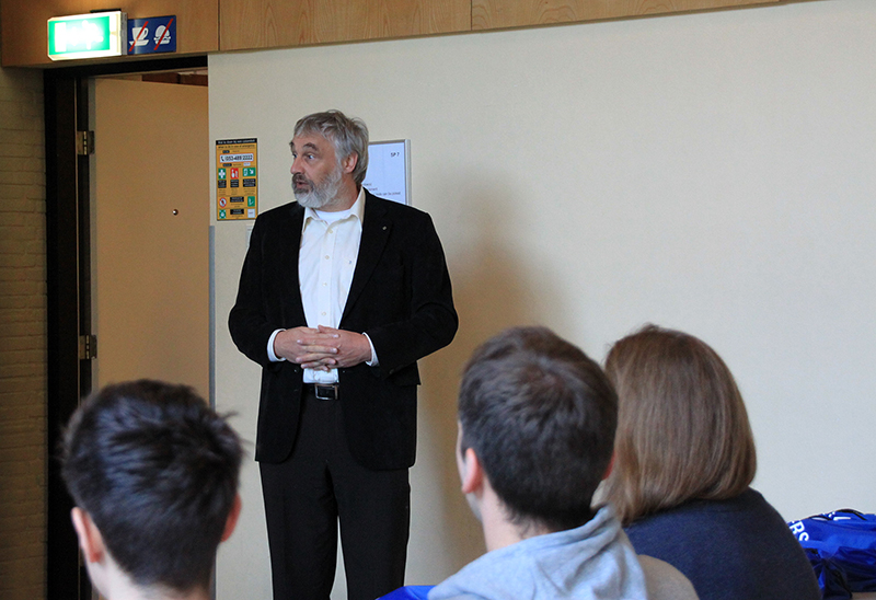 Ein Mann im schwarzen Anzug steht da und spricht zu einem sitzenden Publikum in einem Raum, der wie ein Unterrichts- oder Konferenzraum der Hochschule Coburg aussieht. Der Raum hat beige Wände und eine halb geöffnete Tür mit einem grünen Ausgangsschild darüber, während vier Personen im hinteren Bereich aufmerksam zuhören.