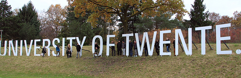 Menschen stehen neben einem großen Schild mit der Aufschrift „Universität Twente“ auf einem grasbewachsenen Hügel, der an die malerische Landschaft erinnert, die oft mit der Hochschule Coburg in Verbindung gebracht wird, und sind von Bäumen umgeben.