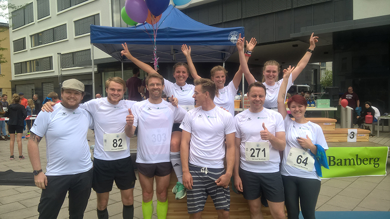 Eine Gruppe von acht Läufern in weißen Shirts der Hochschule Coburg posiert glücklich nach einem Rennen. Sie stehen unter einem blauen Zelt mit Luftballons, einige zeigen mit dem Finger und jubeln. Auf ihren Shirts sind Startnummern zu sehen, im Hintergrund sind ein Gebäude und ein Bamberg-Banner zu sehen.