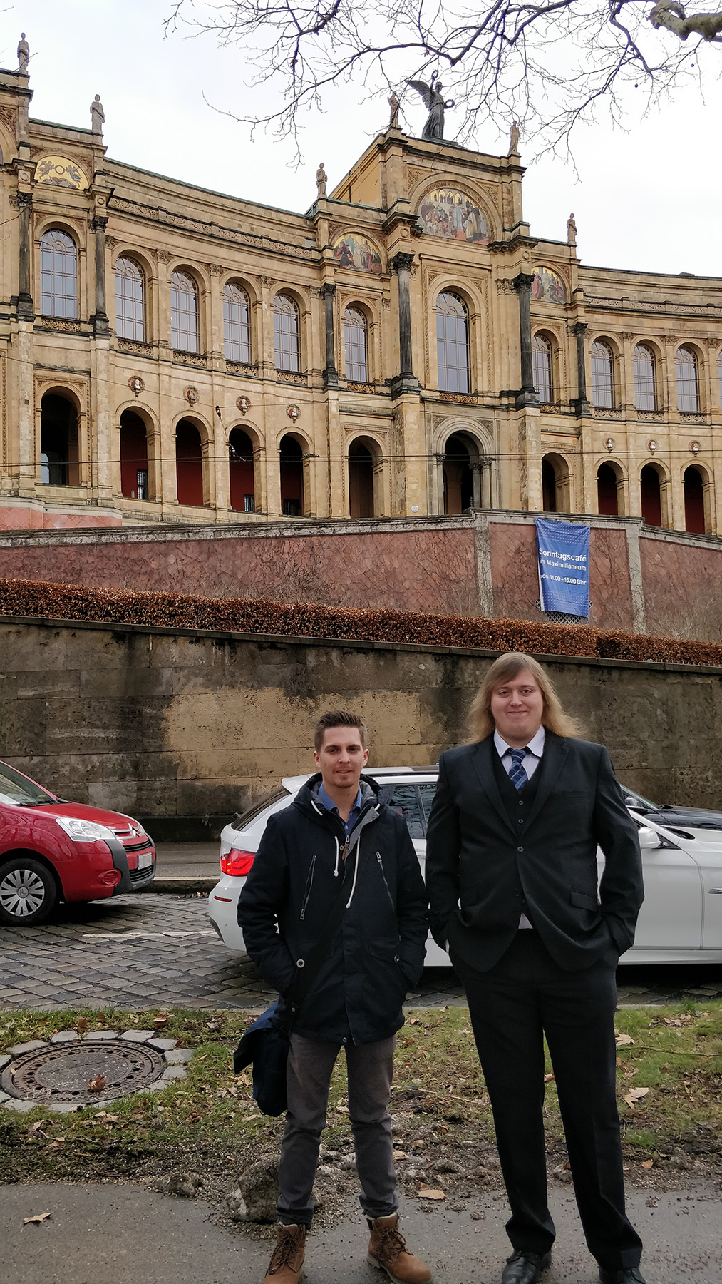 Zwei Personen stehen vor einem kunstvollen, historischen Gebäude mit Bogenfenstern und dekorativem Mauerwerk, das an die Architektur der Hochschule Coburg erinnert. Die Person links trägt eine dunkle Jacke, die Person rechts einen Anzug. Entlang der Straße stehen geparkte Autos.