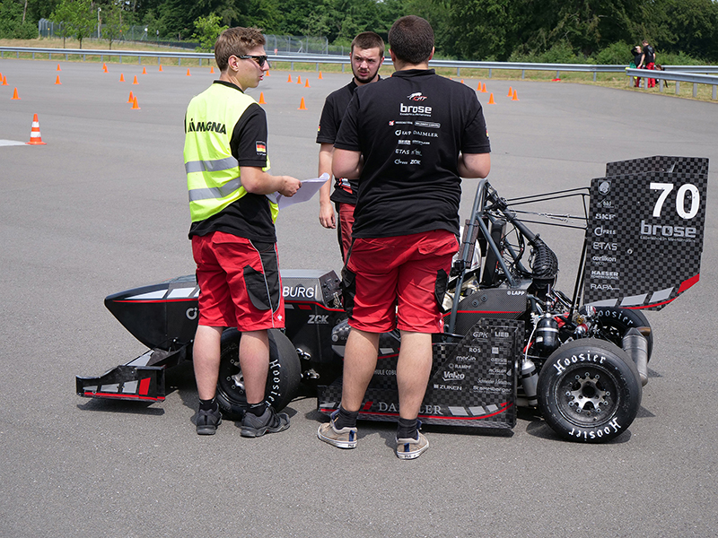 Drei Personen in rot-schwarzen Team-Outfits stehen an der Hochschule Coburg um einen kleinen Open-Wheel-Rennwagen mit der Nummer 70. Eine Person steht in einer Neonweste auf einer Asphaltfläche, im Hintergrund sind orangefarbene Pylonen verteilt.