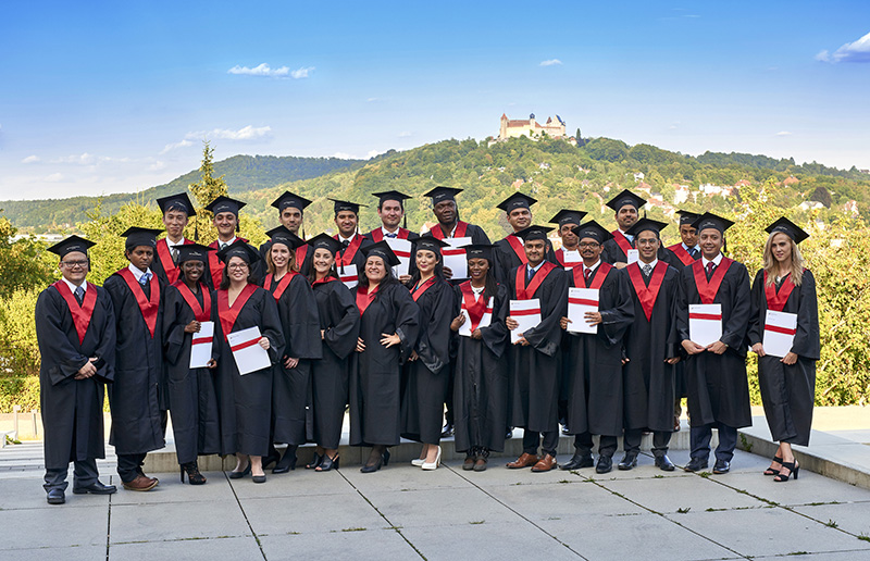 Eine Gruppe von Absolventen der Hochschule Coburg in schwarzen Talaren und Hüten posiert im Freien. Jeder hält ein Diplom in der Hand, während sie auf einer Steinplattform stehen und im Hintergrund einen malerischen Blick auf Hügel und eine Burg unter einem klaren blauen Himmel genießen.