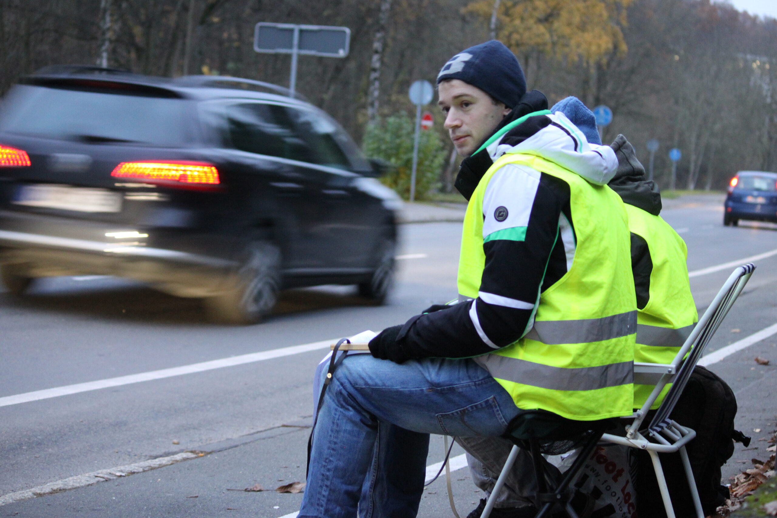 Eine Person in leuchtend gelber Warnweste sitzt auf einem Klappstuhl am Straßenrand in der Nähe der Hochschule Coburg und beobachtet den Verkehr. Die Straße ist von herbstlich gefärbten Bäumen gesäumt. Ein schwarzer SUV fährt vorbei, unscharf durch die Bewegung. Die Atmosphäre ist bewölkt.