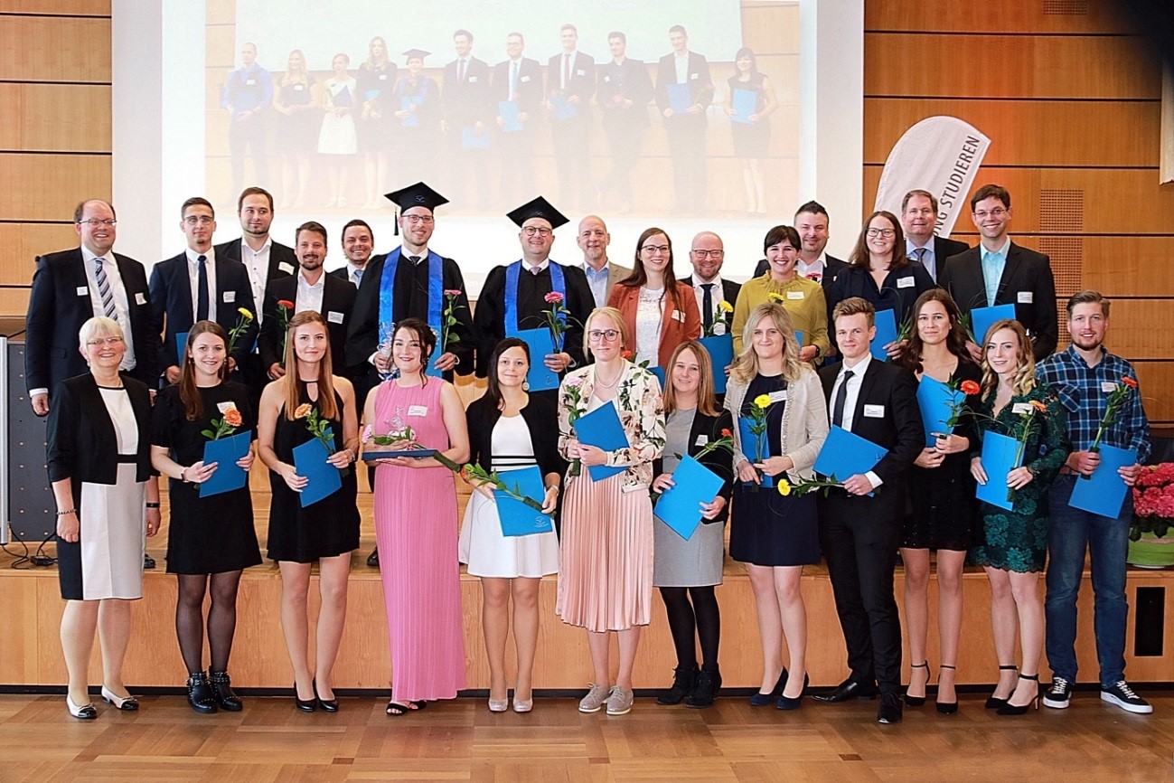 Eine Gruppe von Menschen posiert drinnen, einige tragen Abschlusskappen und Talare und halten stolz Diplome und Blumen in den Händen. Einige Personen sind elegant in Anzügen und Kleidern gekleidet. Hinter ihnen zeigt ein projiziertes Bild ihren denkwürdigen Tag an der Hochschule Coburg.