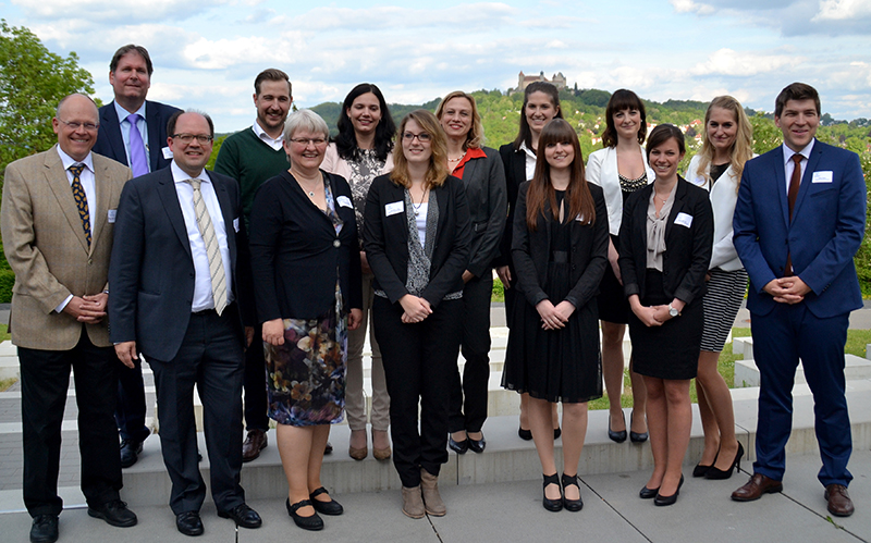 Eine Gruppe von dreizehn Personen, sowohl Männer als auch Frauen, steht im Freien auf einer Terrasse mit malerischer Kulisse. In Business-Kleidung posieren sie für ein Gruppenfoto unter einem teilweise bewölkten Himmel. Das entfernte Gebäude auf dem Hügel hinter ihnen ist Teil des renommierten Campus der Hochschule Coburg.