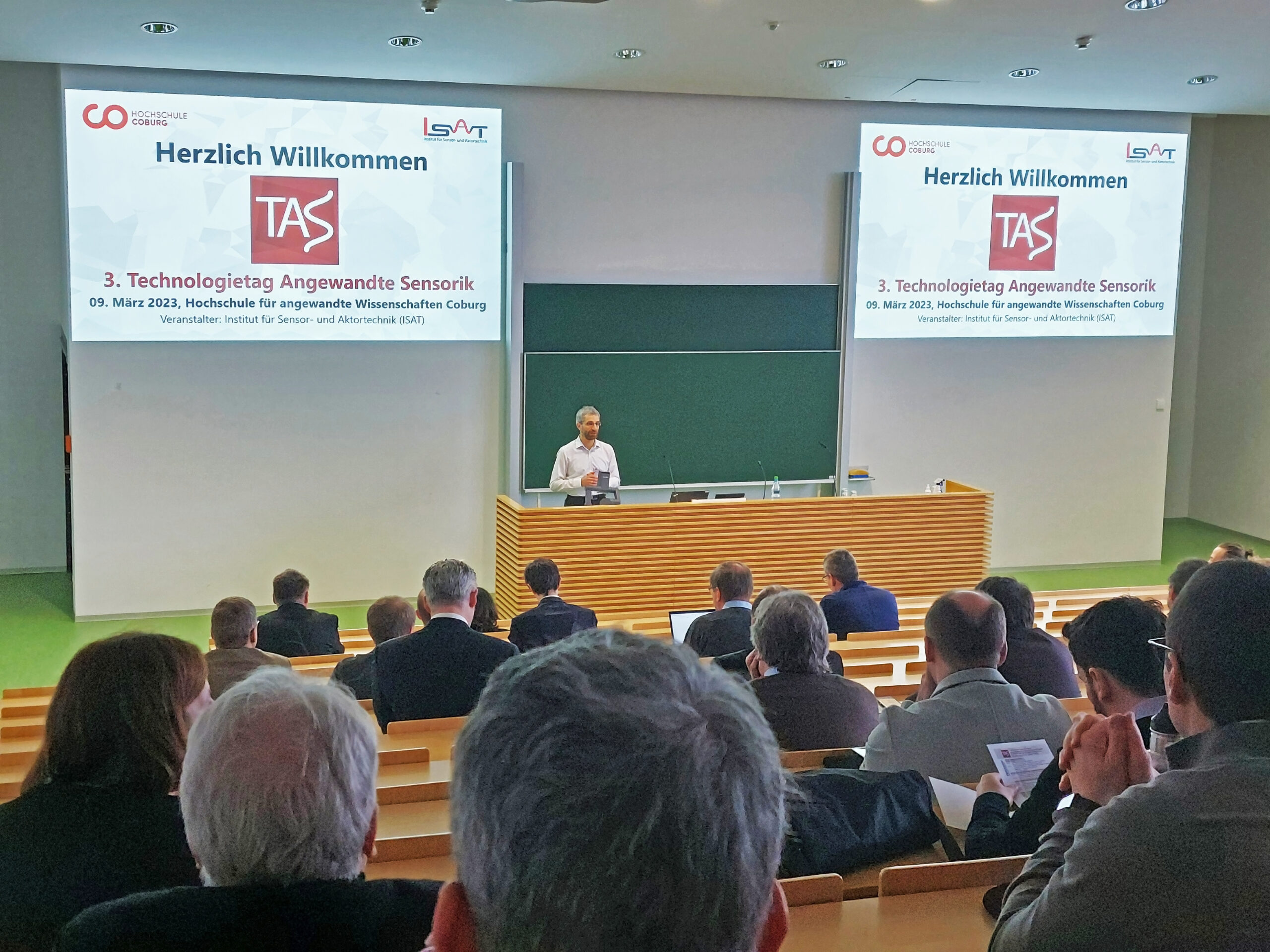 Vor einem Hörsaal mit Holzpult und grüner Tafel hält ein Redner einen Vortrag. Auf zwei großen Bildschirmen sind „Herzlich Willkommen“ und „3. Technologietag Angewandte Sensorik“ an der Hochschule Coburg zu sehen, während ein aufmerksames Publikum in Reihen sitzt.