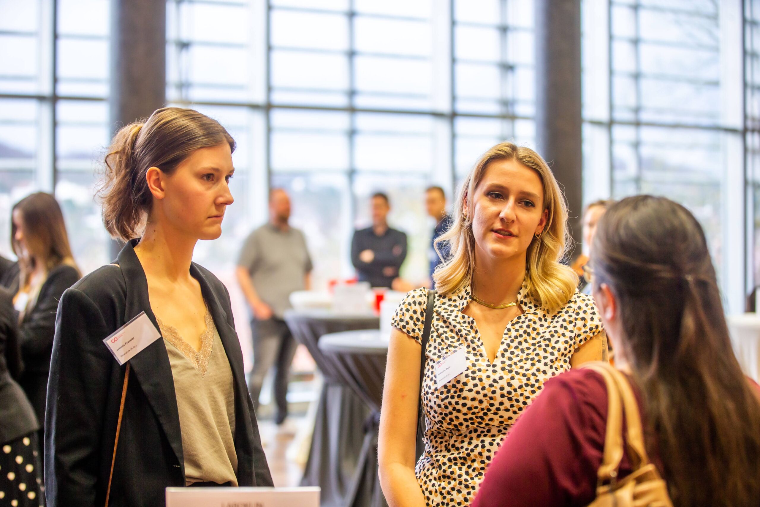 Bei einer Networking-Veranstaltung der Hochschule Coburg gesellt sich eine Gruppe von Menschen zu Gesprächen, wobei zwei Frauen in ein Gespräch vertieft sind. Sie stehen im Innenbereich inmitten anderer und runder Tische im unscharfen Hintergrund. Große Fenster durchfluten den Raum mit natürlichem Licht und schaffen eine einladende Atmosphäre für Kontakte.