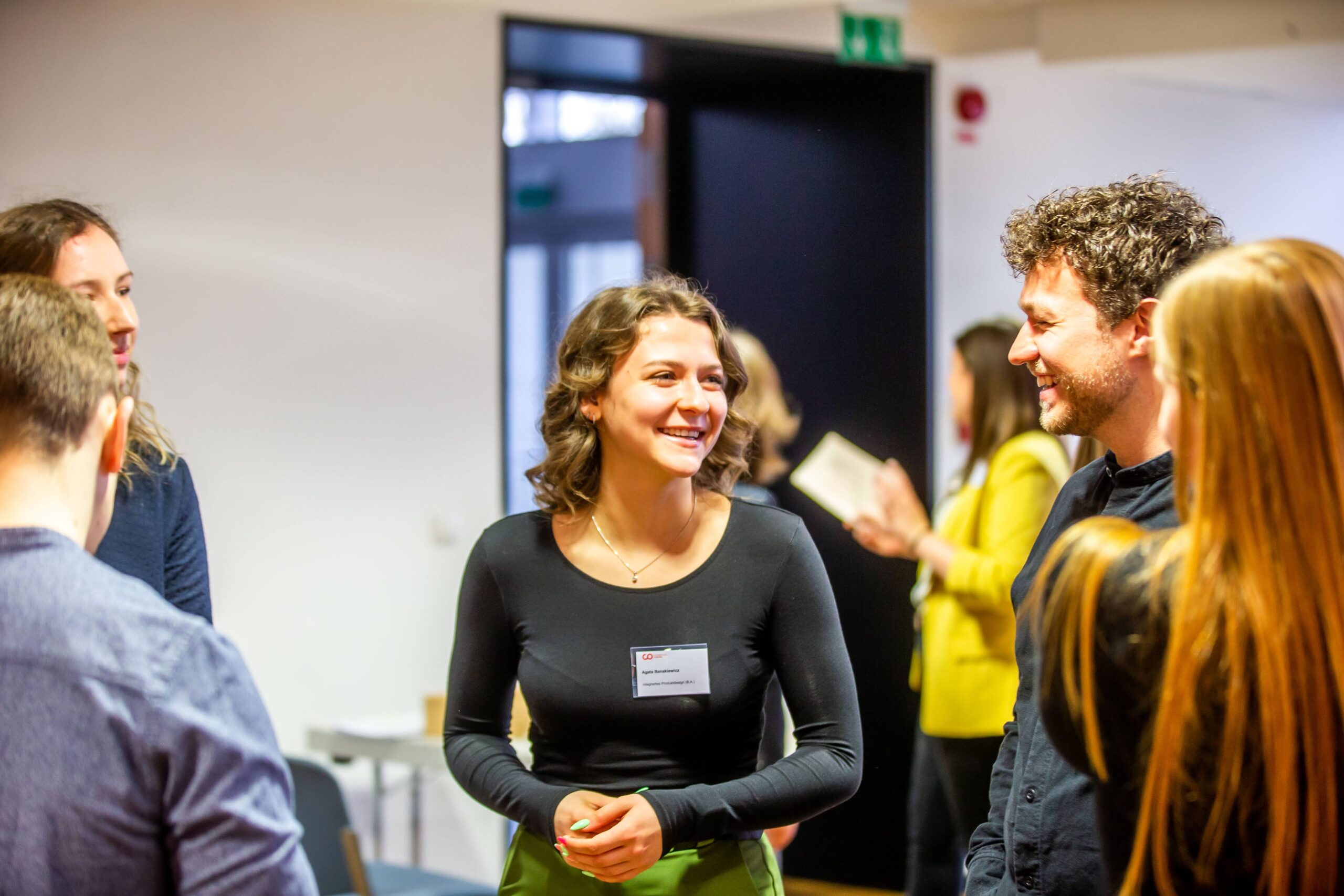 Eine Gruppe von Menschen lächelt und unterhält sich bei einer Indoor-Veranstaltung der Hochschule Coburg. In der Mitte steht eine Frau in einem schwarzen Hemd und unterhält sich mit anderen. Im Hintergrund ist ein offener Raum mit einer Tür und einigen Teilnehmern in der Ferne zu sehen.