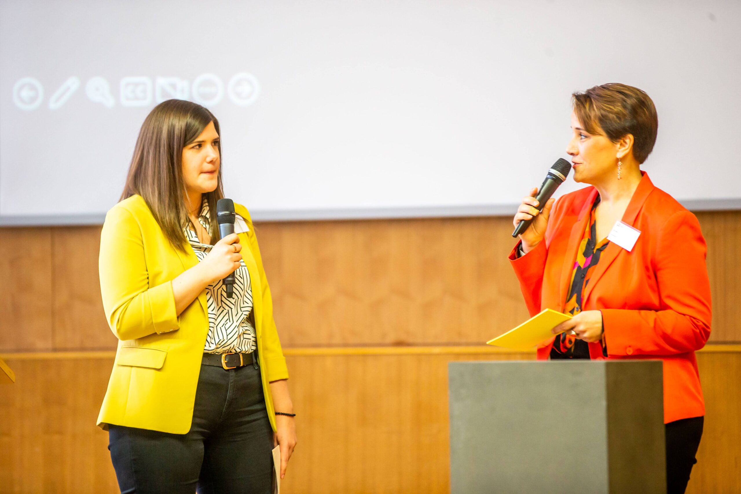 Zwei Frauen stehen mit Mikrofonen auf der Bühne der Hochschule Coburg und unterhalten sich angeregt. Die eine trägt einen gelben Blazer mit gemustertem Hemd, die andere einen roten Blazer. Im Hintergrund ist dezent ein Präsentationsbildschirm zu sehen.