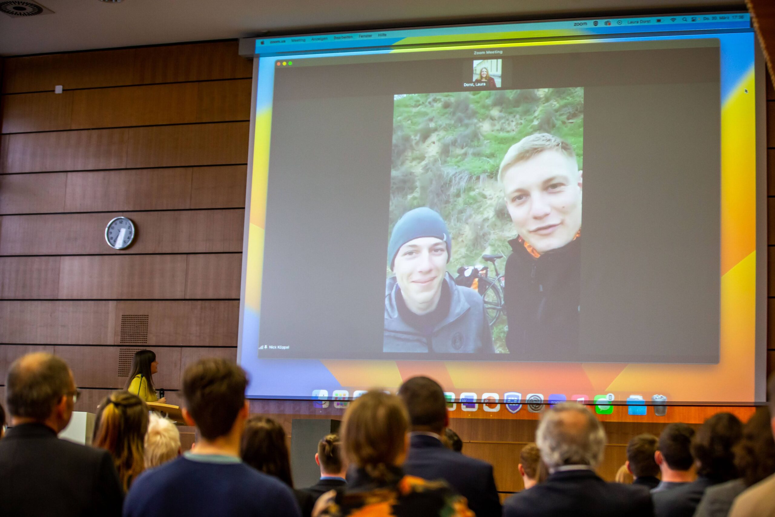 Eine Gruppe von Menschen in einem Konferenzraum der Hochschule Coburg sieht sich das projizierte Bild eines Videoanrufs an, auf dem zwei lächelnde Personen im Freien zu sehen sind. Eine Frau steht vorne am Podium, während eine Uhr an der Wand 10:10 anzeigt.