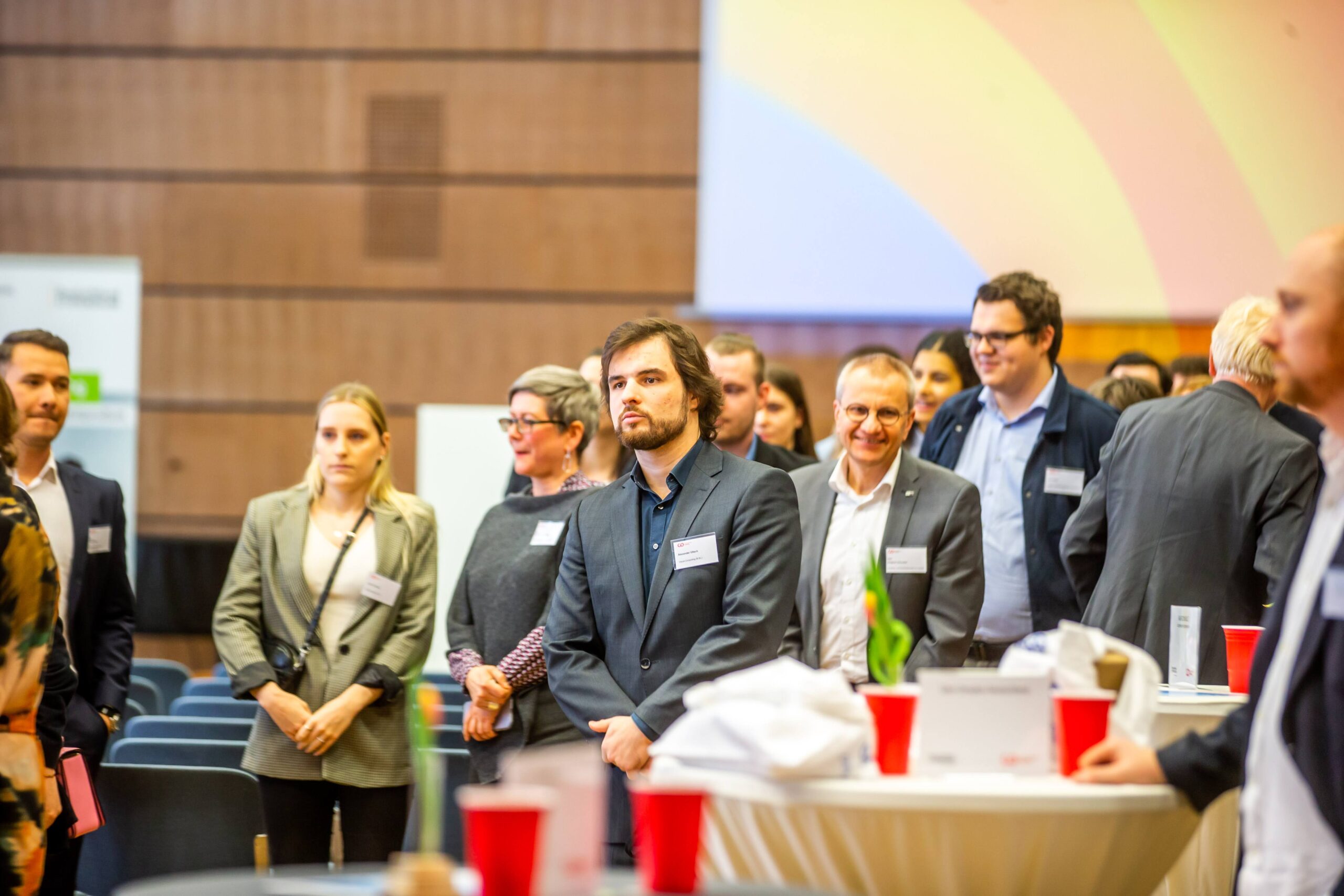 Eine Gruppe von Menschen in Business-Kleidung steht in einem Konferenzsaal der Hochschule Coburg und wirkt aufmerksam. Tische mit roten Bechern und Papieren sind zu sehen, und ein großer Bildschirm zeigt einen Farbverlauf-Hintergrund. Die formelle Umgebung konzentriert sich auf eine Präsentation oder Rede.