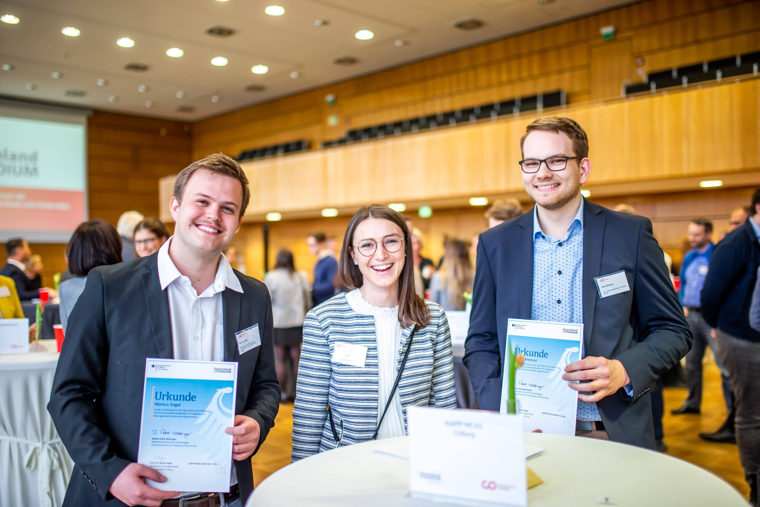 Drei lächelnde Personen halten Zertifikate bei einer Indoor-Veranstaltung der Hochschule Coburg. Sie stehen um einen Tisch im Konferenzsaal, im Hintergrund sind unscharfe Teilnehmer zu sehen. Die Umgebung wirkt professionell und gut beleuchtet.