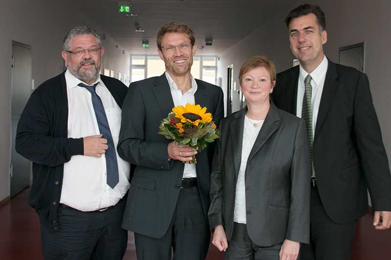 Vier Personen stehen in einem Flur der Hochschule Coburg. Drei Männer und eine Frau sind in Business-Kleidung gekleidet und strahlen eine formelle Atmosphäre aus. Ein Mann hält einen Blumenstrauß mit einer Sonnenblume in der Hand und alle lächeln herzlich.