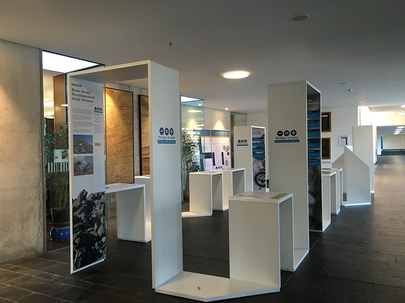 Der moderne Ausstellungsbereich der Hochschule Coburg besteht aus weißen Ausstellungsständen, die in einem geräumigen Flur angeordnet sind. Jeder Stand präsentiert Texte und Bilder zu Umweltthemen. Der Raum ist mit natürlichem und künstlichem Licht gut beleuchtet, während Pflanzen die Ecken schön schmücken.