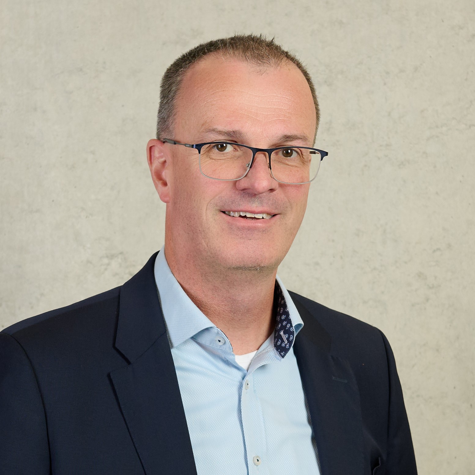 Ein lächelnder Mann mit kurzen dunklen Haaren und Brille trägt ein weißes Hemd und einen marineblauen Blazer. Er steht stolz vor einem farbenfrohen abstrakten Hintergrund in Rot-, Blau- und Cremetönen und verkörpert den lebendigen Geist der Hochschule Coburg.