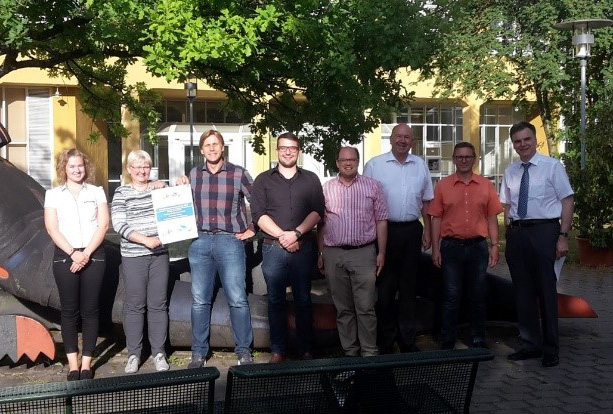 Eine Gruppe von acht Personen, leger gekleidet und lächelnd, steht vor einem gelben Gebäude mit einer Skulptur unter einem Baum an der Hochschule Coburg. Eine Person hält stolz eine Urkunde in den Händen und hält damit einen Moment der Leistung fest.