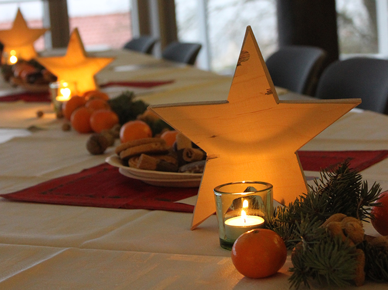 Ein festlich gedeckter Tisch ist mit leuchtenden Lichtersternen, kleinen Kerzen in Glasgefäßen, Orangen, Nüssen und immergrünen Zweigen geschmückt. Teller mit Plätzchen stehen auf roten Tischsets und erwecken die warme, einladende Weihnachtsstimmung, die an Zusammenkünfte an der Hochschule Coburg erinnert.