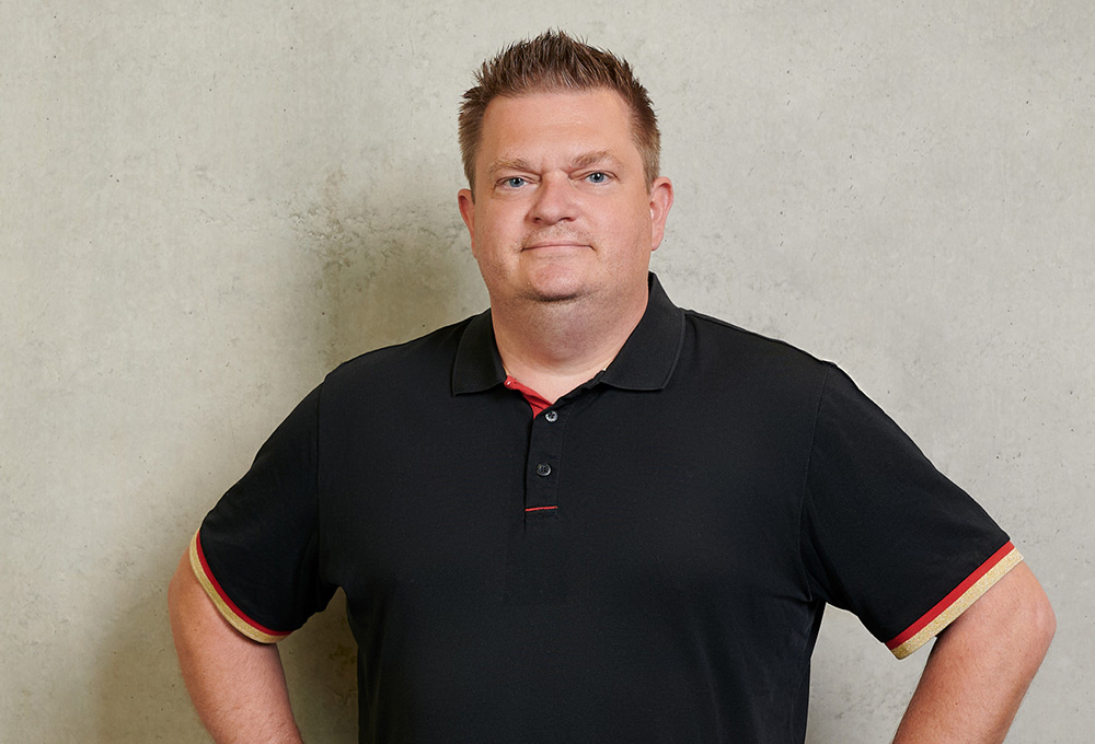 A man with short hair stands against a plain, light-colored wall, proudly showcasing his Hochschule Coburg polo shirt. The black fabric is accented with red and gold, perfectly coordinating as he confidently rests his hands on his hips.