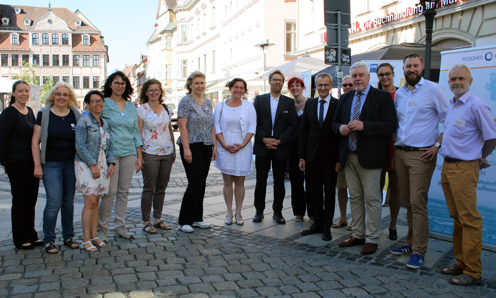 Eine Gruppe von Menschen steht zusammen auf einer Kopfsteinpflasterstraße in einem sonnigen Stadtgebiet in der Nähe der Hochschule Coburg. Einige tragen formelle Kleidung, andere tragen Freizeitkleidung. Im Hintergrund sind Gebäude und ein Banner mit Text zu sehen.