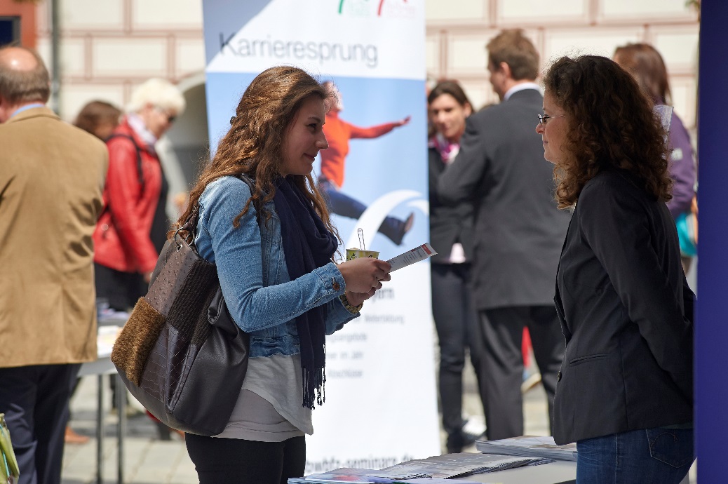 Zwei Frauen unterhalten sich auf einer Outdoor-Karrieremesse der Hochschule Coburg vor einem Stand mit der Aufschrift „Karrieresprung“. Im Hintergrund wimmelt es von mehreren Menschen und bunten Bannern, was für eine lebhafte Atmosphäre sorgt.