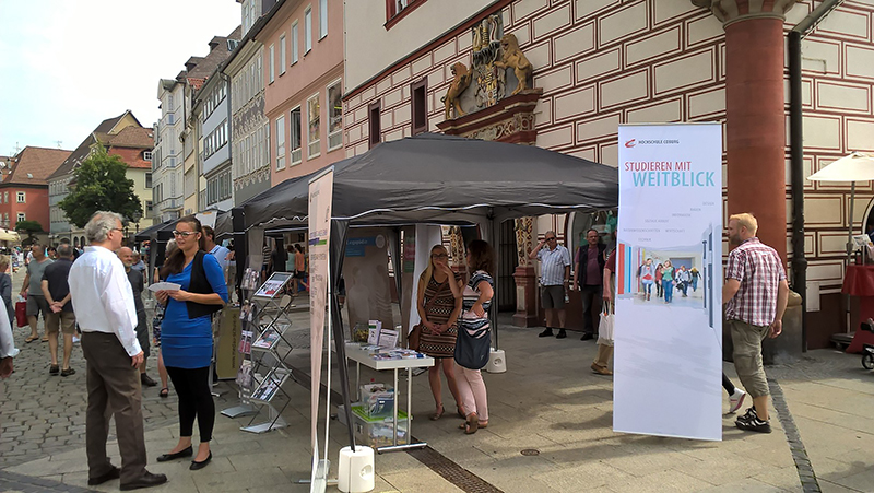 Auf einem lebhaften Straßenfest steht ein Zelt mit einem Informationsstand der Hochschule Coburg. Menschen versammeln sich um das Zelt und stöbern in Materialien auf Tischen unter einem Banner am Eingang, während im Hintergrund Gebäude aufragen und der lebhaften Szene einen akademischen Touch verleihen.