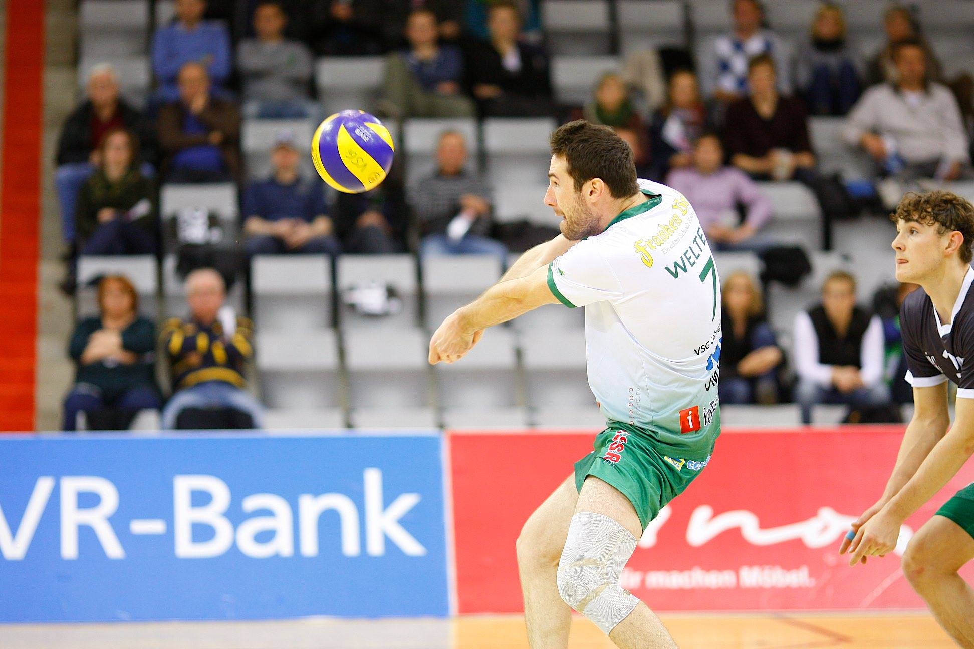 Ein Volleyballspieler der Hochschule Coburg in weiß-grüner Uniform stürzt sich auf einen Ball in der Luft, während ein anderer Spieler in schwarz-weißer Uniform daneben steht. Im Hintergrund verfolgen die Zuschauer gespannt das spannende Spiel.