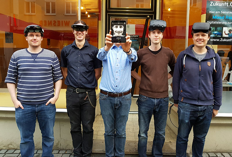 Fünf Personen stehen vor einem Gebäude, vier tragen VR-Headsets. Die Person in der Mitte hält ein Tablet, auf dessen Bildschirm ein Gesicht angezeigt wird. Sie sind leger gekleidet und scheinen an einer Technologiedemonstration an der Hochschule Coburg teilzunehmen.