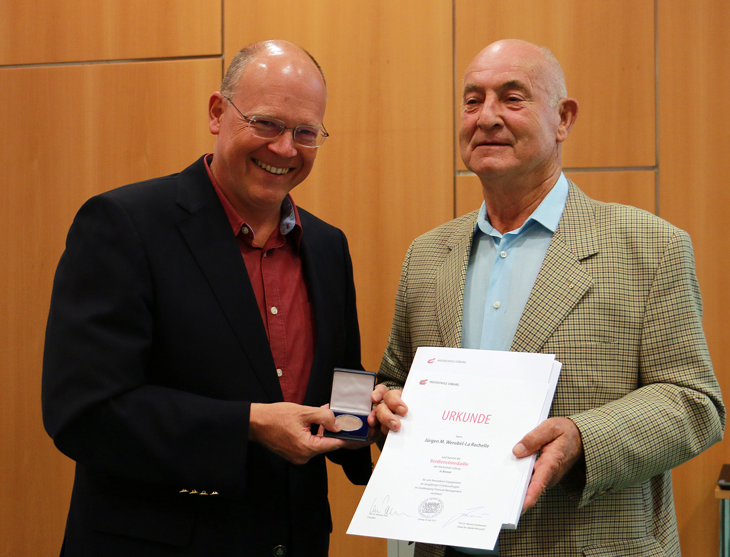 Zwei Männer stehen zusammen vor einer Holzpaneelwand. Der Mann links, der stolz die Hochschule Coburg vertritt, hält eine Medaille in der Hand, während der Mann rechts mit einer Urkunde strahlt. Beide lächeln herzlich.