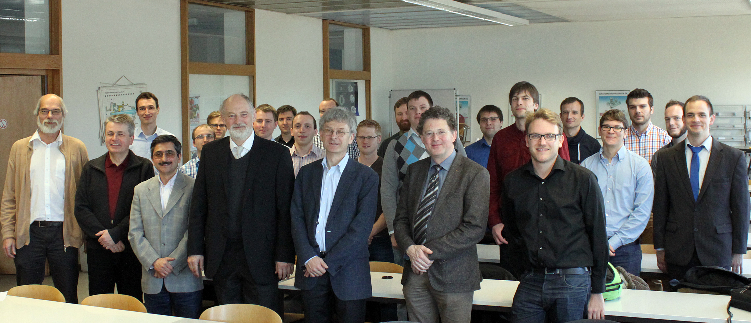 Eine Gruppe von Männern posiert für ein Foto in einem modernen Konferenzraum der Hochschule Coburg. Sie sind in Business- und Freizeitkleidung gekleidet und stehen hinter einer Reihe von Schreibtischen mit großen Fenstern und Postern an den Wänden, die zur zeitgenössischen Ästhetik beitragen.