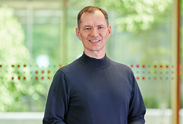 Eine Person mit kurzen Haaren steht lächelnd im Innenbereich der Hochschule Coburg. Sie trägt ein dunkles Langarmshirt und im Hintergrund ist ein großes Fenster mit Blick auf das üppige grüne Laub draußen zu sehen.