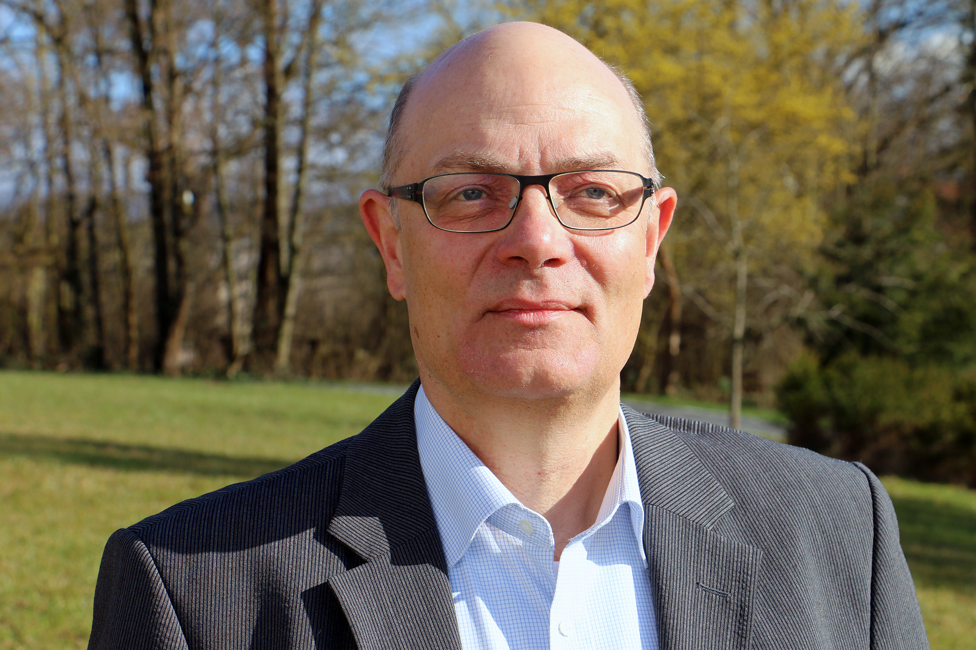 Ein Mann mit Brille und Anzug steht mit ruhigem Gesichtsausdruck im Freien in der Nähe der Hochschule Coburg. Er befindet sich auf einer Wiese mit Bäumen im Hintergrund, unter einem klaren Himmel.