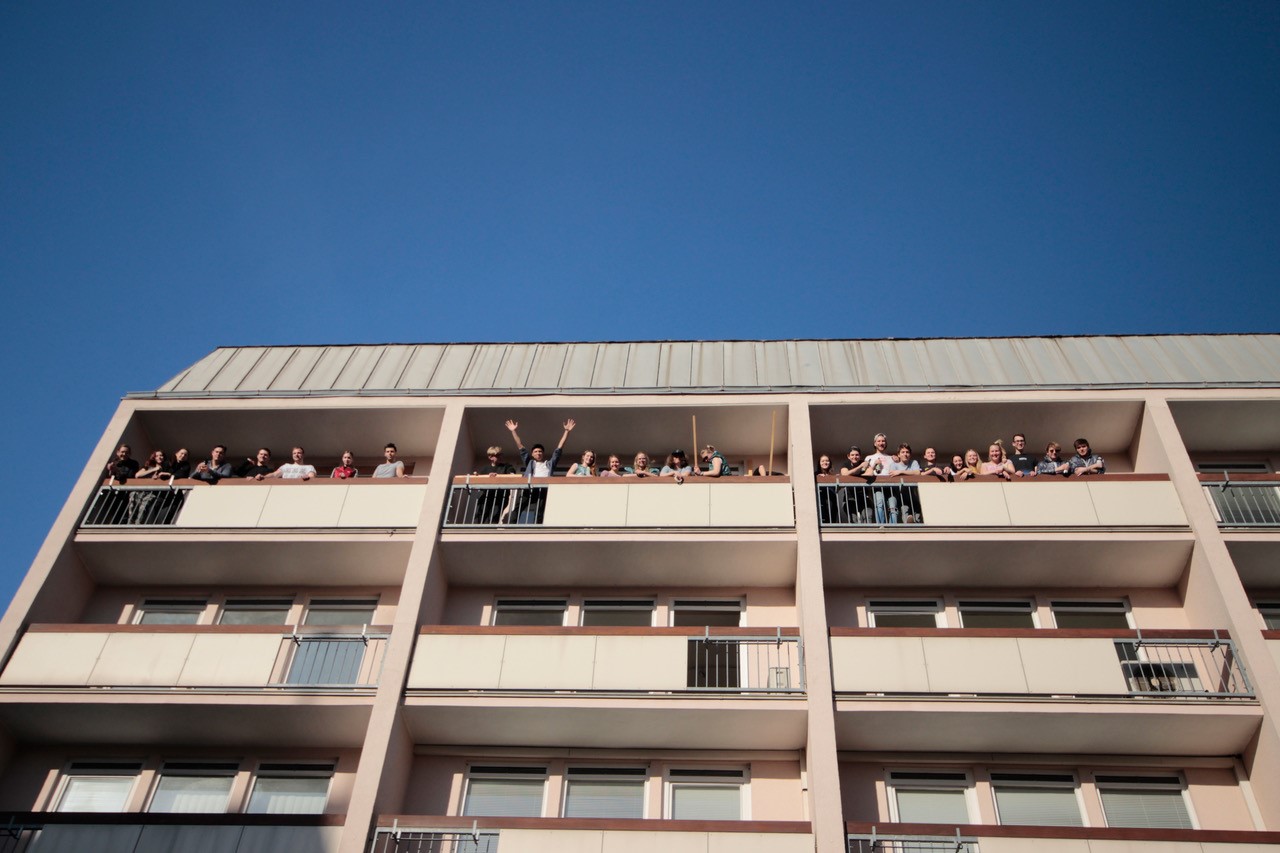 Menschen stehen auf den Balkonen eines beigefarbenen, mehrstöckigen Wohnhauses in der Nähe der Hochschule Coburg und blicken in den klaren blauen Himmel. Einige winken oder heben zur Begrüßung die Hand.