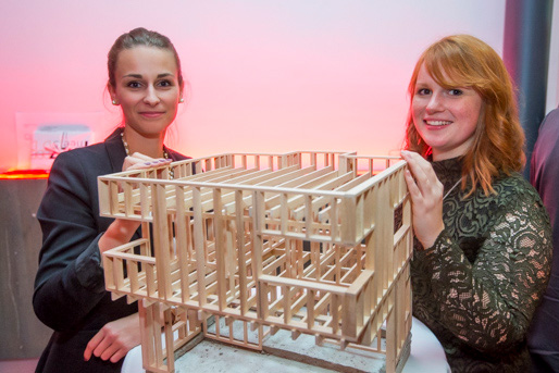 Zwei Frauen der Hochschule Coburg lächeln und halten ein hölzernes Architekturmodell eines mehrstöckigen Gebäudes in der Hand. Der Hintergrund ist sanft in einem rosa Farbton beleuchtet. Sie sind in formelle Kleidung gekleidet und scheinen sich auf einer Veranstaltung oder Ausstellung zu befinden.