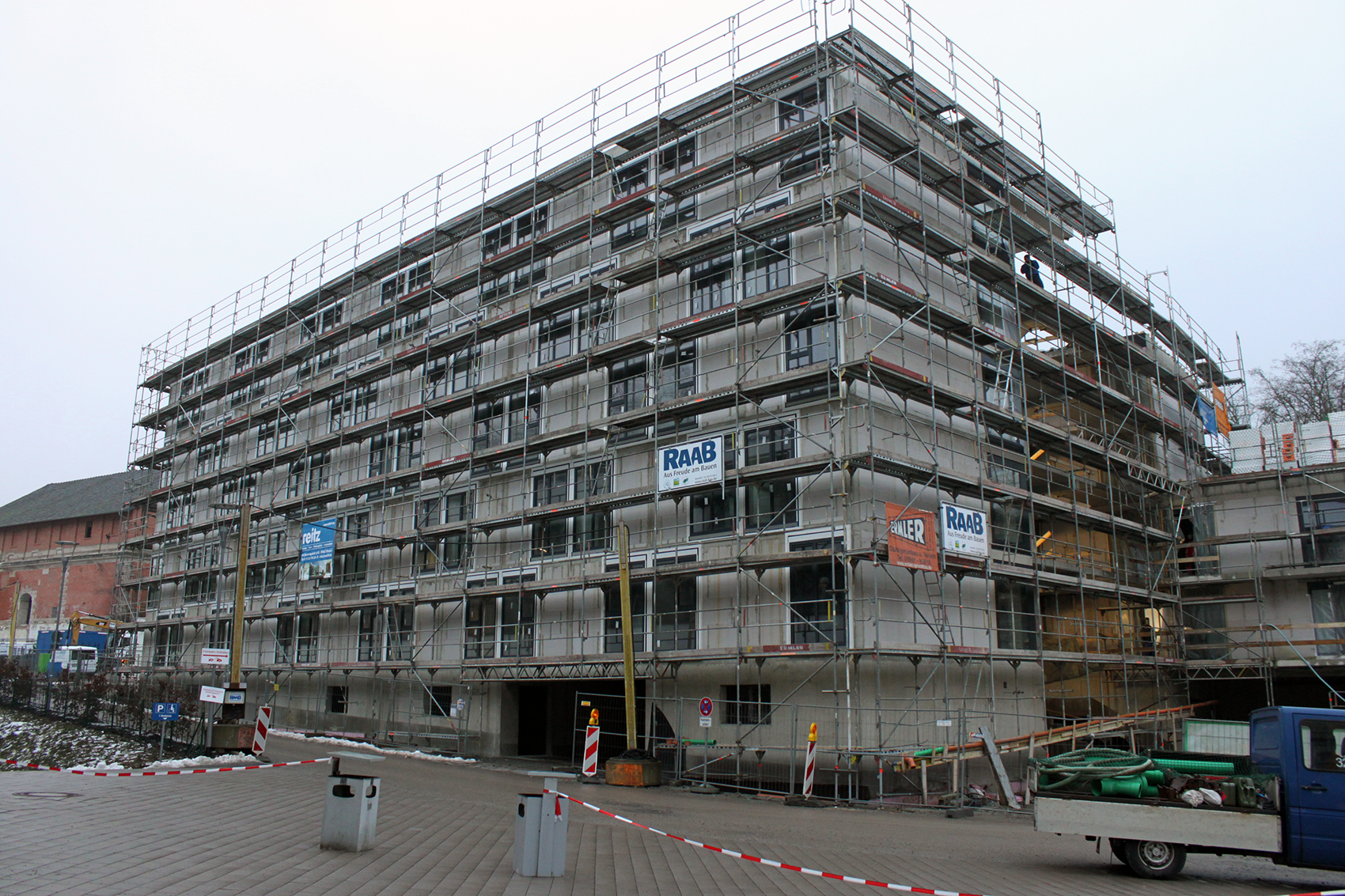 Ein im Bau befindliches mehrstöckiges Gebäude der Hochschule Coburg ist von Gerüsten umgeben. Im Vordergrund sind Absperrungen und Schilder zu sehen. In der Nähe parkt ein LKW, was auf die laufenden Bauarbeiten auf der geschäftigen Baustelle hinweist.