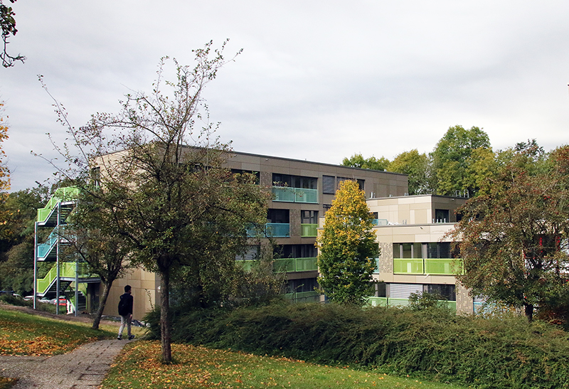 Ein moderner Apartmentkomplex der Hochschule Coburg ist von Grün umgeben, im Vordergrund sind Bäume und Sträucher zu sehen. Links geht eine Person einen Weg entlang. Der bewölkte Himmel wirft ein neutrales Licht auf die Szene und schafft eine ruhige Atmosphäre in der Nähe des akademischen Zentrums.