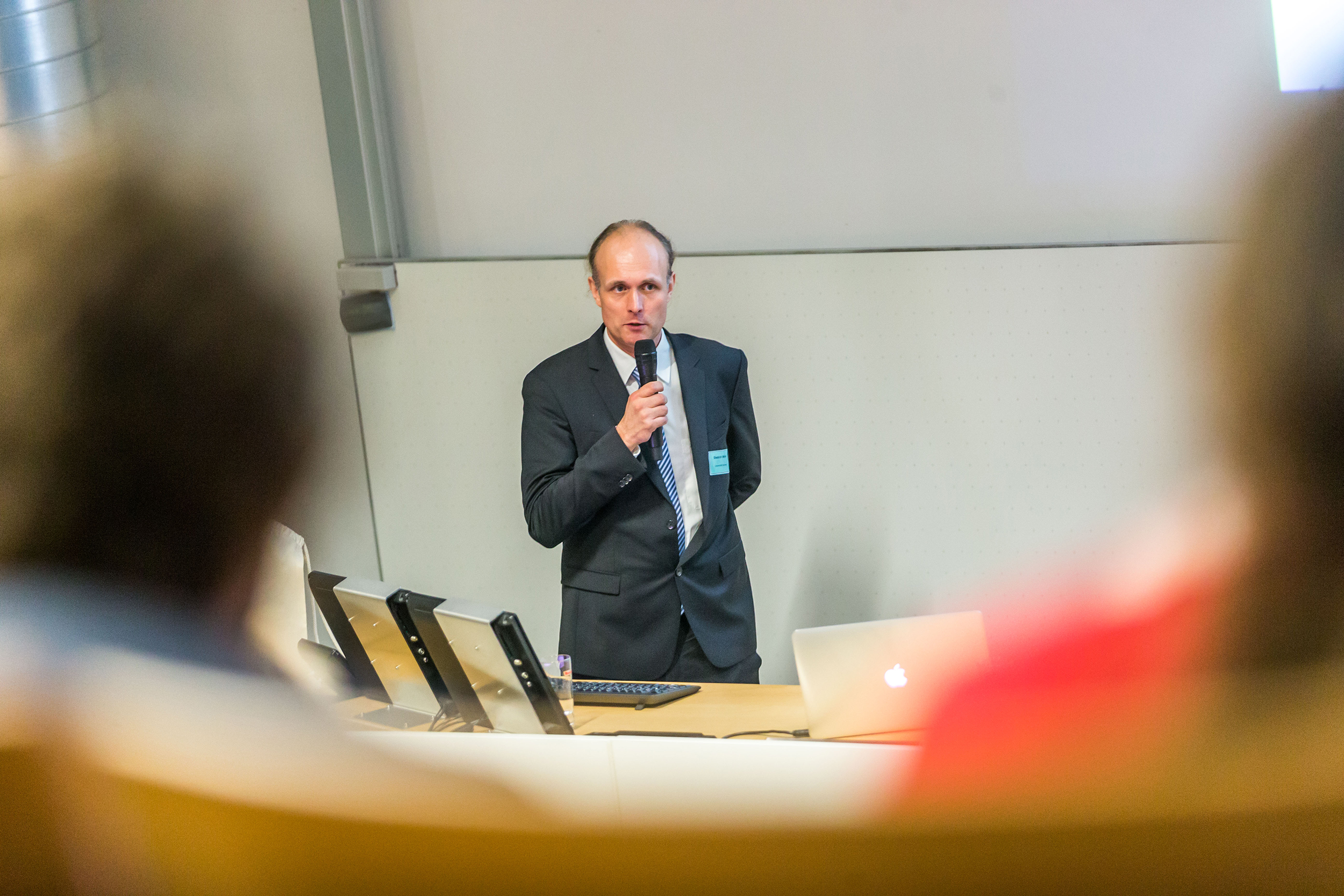 Eine Person im Anzug steht vorne im Hörsaal der Hochschule Coburg und spricht in ein Mikrofon. Hinter einem Rednerpult mit einem Laptop wendet sie sich an die Teilnehmer, die im Vordergrund sitzen und alle diesem engagierten Redner zugewandt sind.