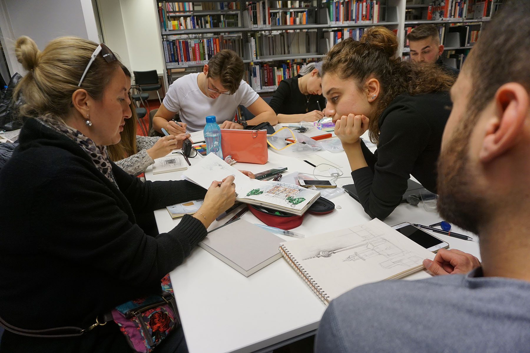 Eine Gruppe von Menschen, möglicherweise von der Hochschule Coburg, sitzt in einer Bibliothek um einen Tisch und beschäftigt sich mit Zeichenaktivitäten. Der Tisch ist mit Skizzenbüchern, Bleistiften und Künstlerbedarf gefüllt, während Bücherregale voller Bände eine inspirierende Kulisse bilden.