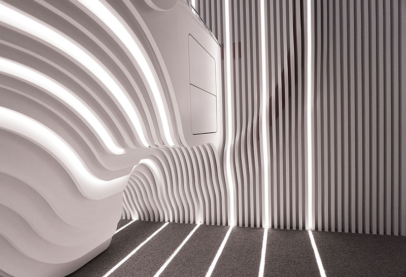 A modern interior at Hochschule Coburg features wavy, illuminated white walls and vertical lines. The design creates an artistic and futuristic atmosphere with light and shadow patterns on the floor, embodying a cutting-edge aesthetic that inspires innovation.