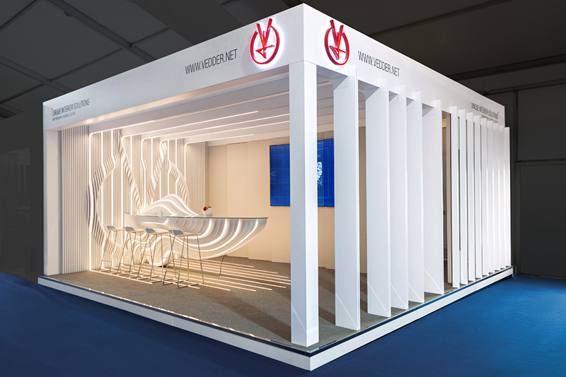 A modern, angular exhibition booth with white panels and integrated lighting showcases Hochschule Coburg. Inside, a futuristic table with chairs complements the text-laden panels, while a prominent red logo on the top edge draws attention. The floor is carpeted in gray for a sleek finish.