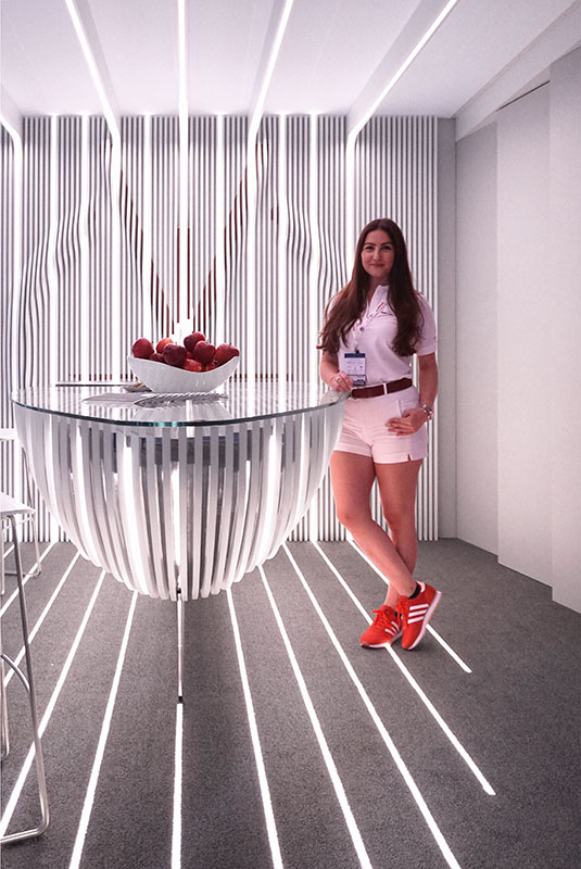 A woman stands beside a modern, striped counter at Hochschule Coburg with a bowl of red apples on top. She wears a white shirt, pink shorts, and red sneakers in the futuristic room, where light streams in from above.