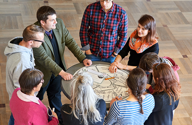 Eine Gruppe von acht Personen steht an einem runden Tisch in der Hochschule Coburg und diskutiert. Auf dem Tisch ist ein Diagramm mit Text und Abbildungen zu sehen. Die Kulisse scheint ein geräumiger Raum mit Holzboden zu sein.