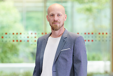 Ein kahlköpfiger Mann mit Bart steht selbstbewusst in einem Innenraum der Hochschule Coburg. Er trägt ein weißes Hemd und einen grauen Blazer, während große Fenster hinter ihm den Blick auf das üppige Grün draußen freigeben.