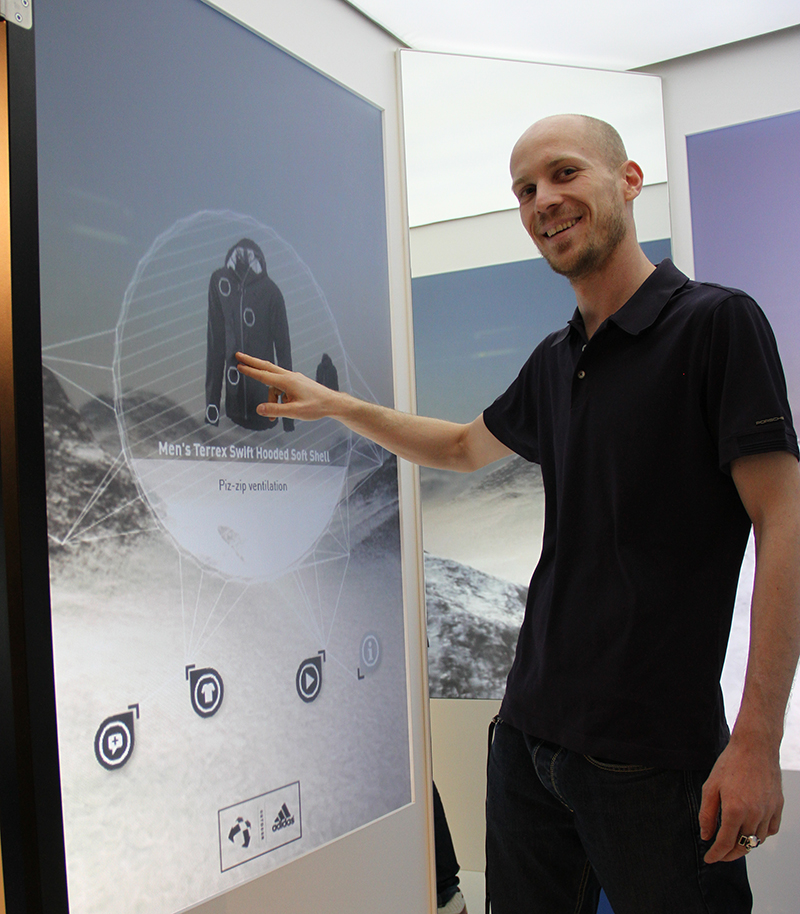 Eine Person in einem schwarzen Poloshirt bedient ein großes digitales Touchscreen-Display, auf dem eine Terrex Swift Hooded Softshell-Jacke für Herren vor der malerischen Berglandschaft der Hochschule Coburg im Hintergrund präsentiert wird.